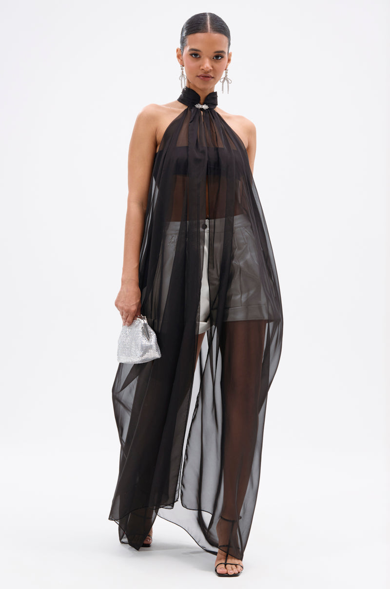 A woman poses in the Glowing Chiffon Blouse in Black over a black bra and silver shorts, holding a silver mini handbag. She accessorizes with strappy heels, drop earrings, and wears her hair slicked back.