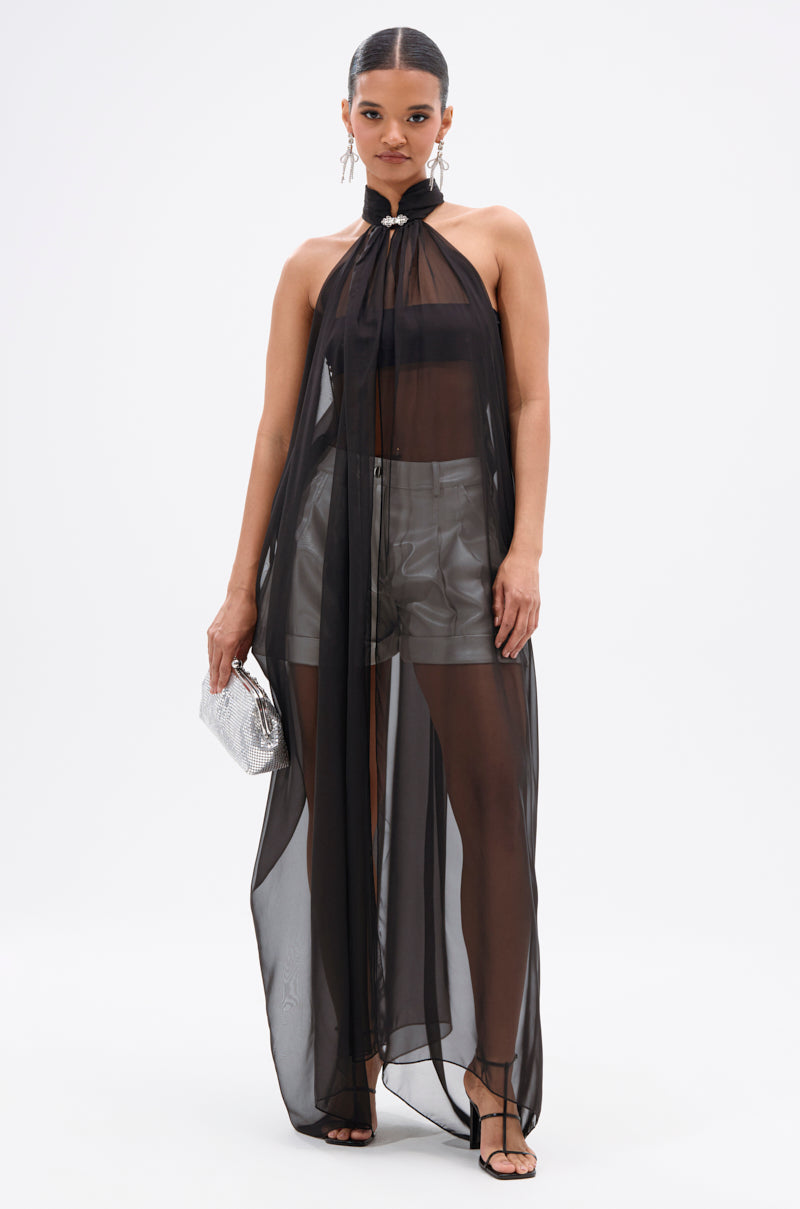A woman poses against a white background wearing the Glowing Chiffon Blouse in Black over a black bralette and gray shorts, styled with statement earrings, a rhinestone silver clutch, and black strappy heels.
