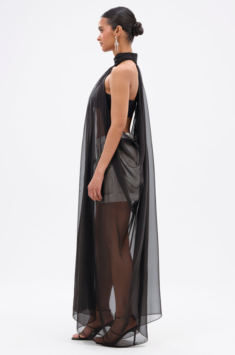 A woman stands in profile wearing the Glowing Chiffon Blouse in Black over a sleeveless mini dress. Her hair is styled in a sleek bun and she accessorizes with strappy heels and long drop earrings, set against a plain white background.