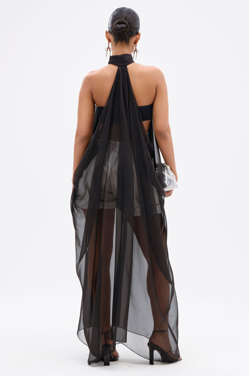 A woman faces away wearing the GLOWING CHIFFON BLOUSE IN BLACK over black shorts and a bandeau top, paired with a small silver clutch, black heels, and her hair in a low bun.