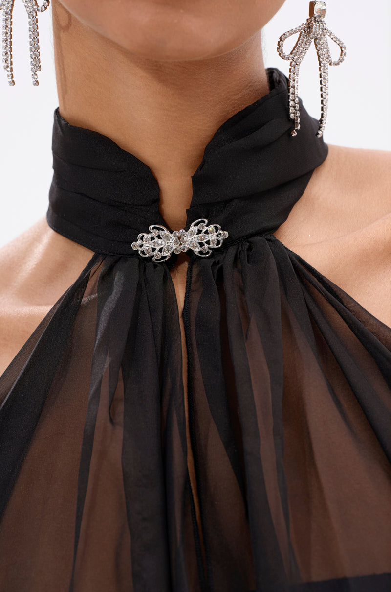 Close-up of a person wearing the GLOWING CHIFFON BLOUSE IN BLACK, a sheer halter-neck top with a high collar fastened by a rhinestone brooch, paired with dangling bow-shaped rhinestone earrings.