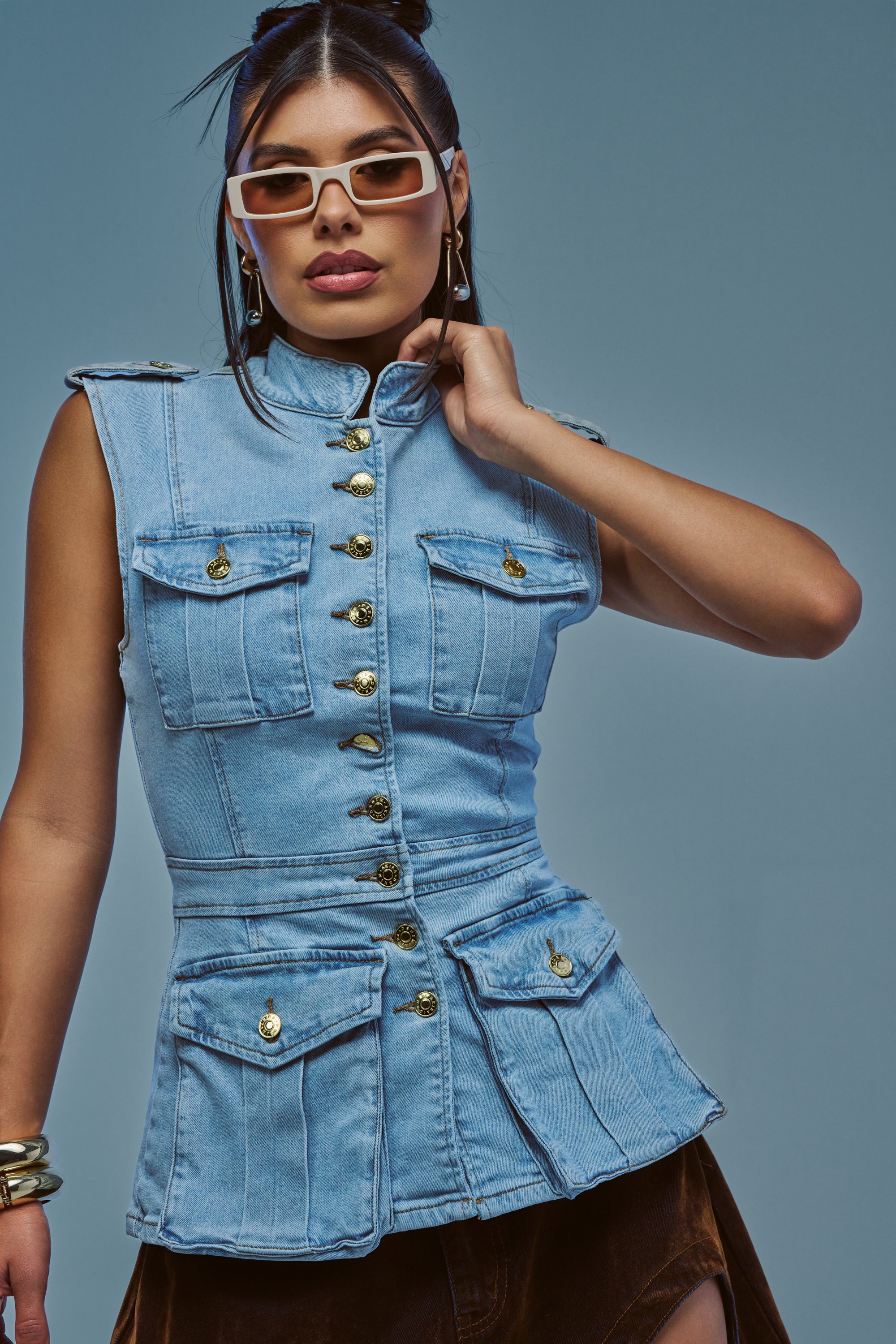 Wearing the UMI Stretch Denim Top in Light Blue Denim, a woman poses against a plain blue background with white sunglasses and gold buttons accenting her sleeveless look, perfectly embodying the utility-chic vibe.