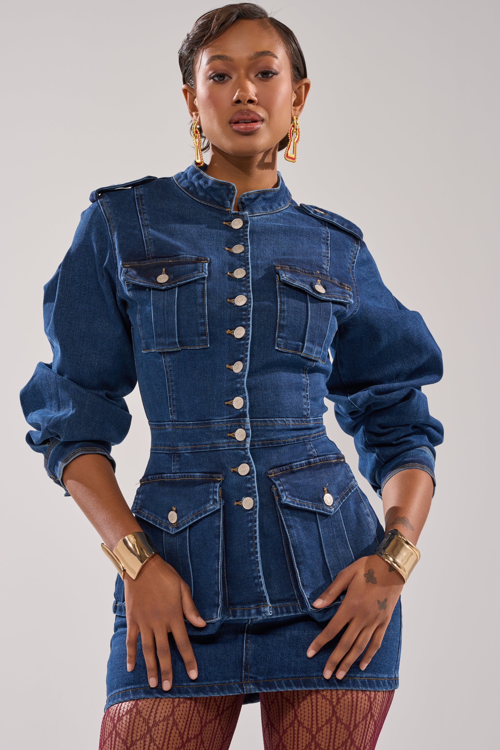 A person poses confidently in the CRAZY FOR YOU LONG SLEEVE DENIM BUTTON DOWN styled as a jacket-dress with silver buttons and multiple pockets, accessorized with gold cuff bracelets and geometric earrings against a plain light background.