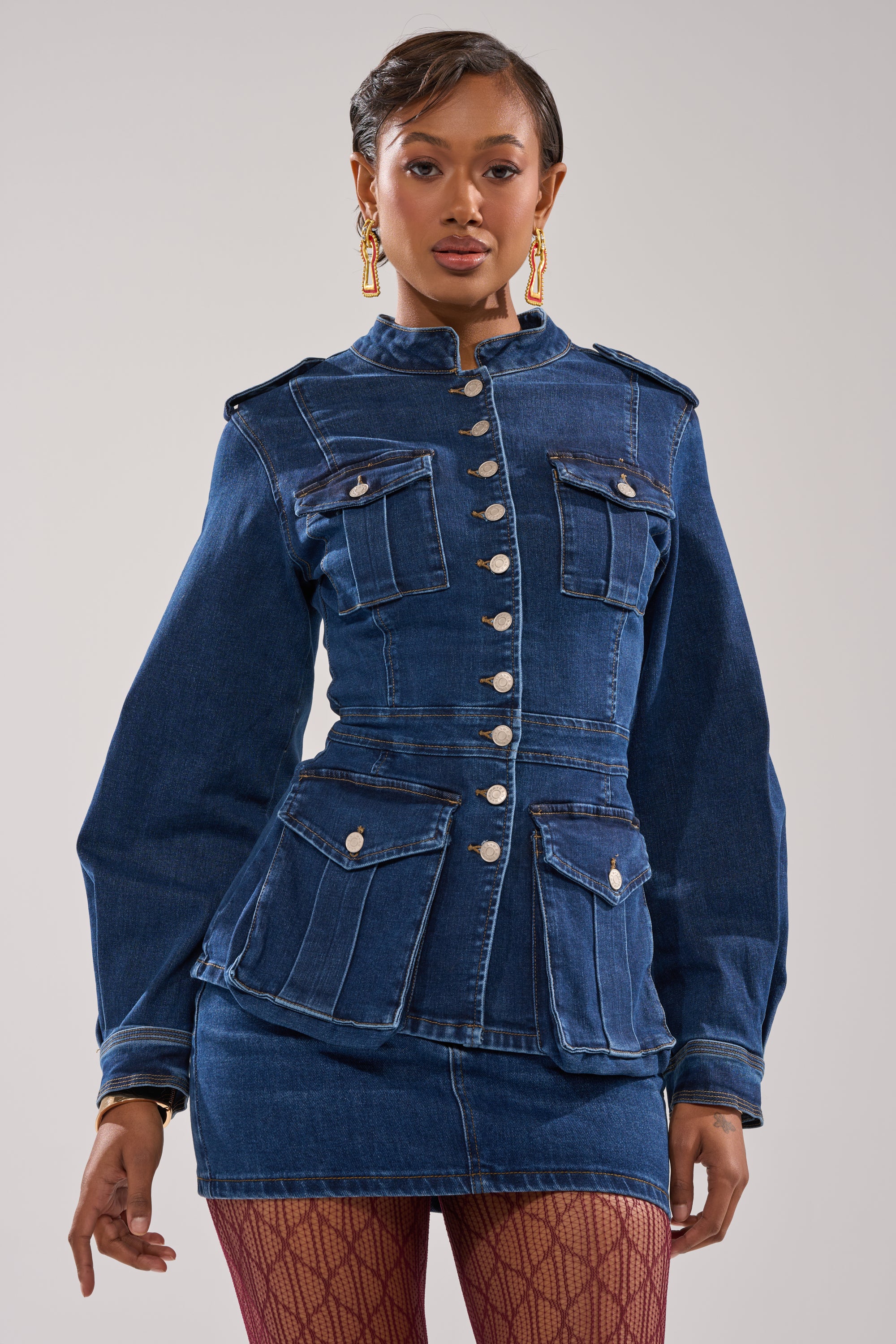 A woman poses confidently in the CRAZY FOR YOU LONG SLEEVE DENIM BUTTON DOWN with silver buttons, a matching denim mini skirt, patterned red tights, and gold earrings against a light gray background.