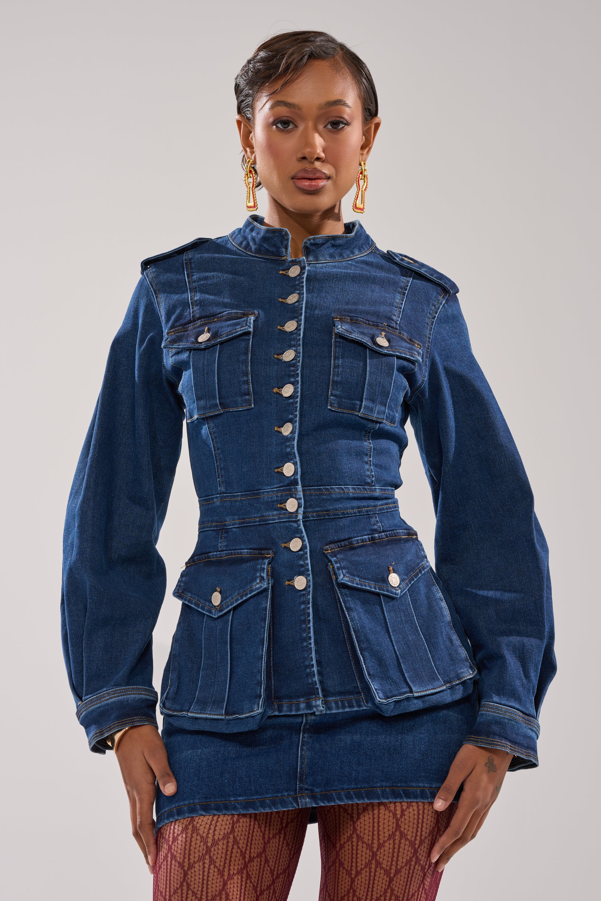 A woman poses confidently in the CRAZY FOR YOU LONG SLEEVE DENIM BUTTON DOWN with matching mini skirt, gold earrings, and patterned red tights against a plain background.