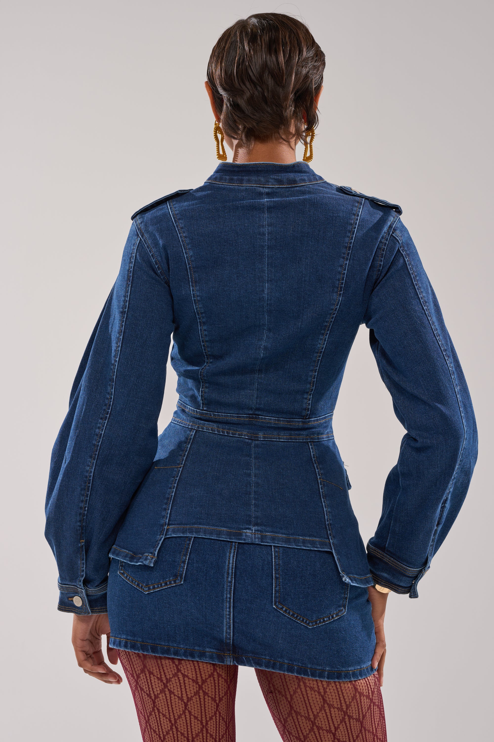 A person with short hair and gold hoop earrings faces away, wearing the CRAZY FOR YOU LONG SLEEVE DENIM BUTTON DOWN with a belt and puffed sleeves, matched with a denim skirt and patterned red tights.