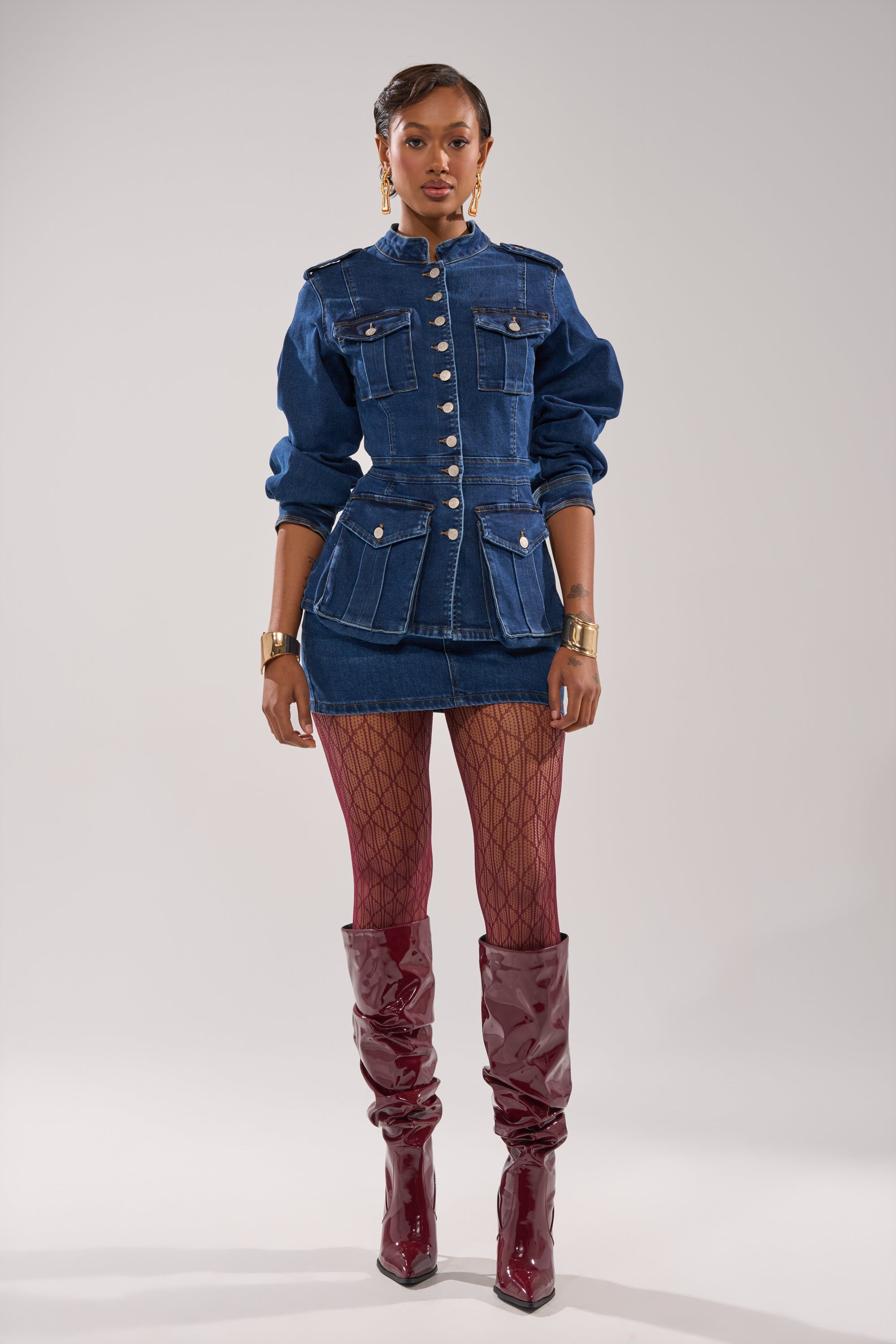 A woman faces forward, wearing the CRAZY FOR YOU LONG SLEEVE DENIM BUTTON DOWN as a jacket over a mini skirt, with red fishnet stockings, burgundy slouchy boots, and gold jewelry, set against a plain light gray background.