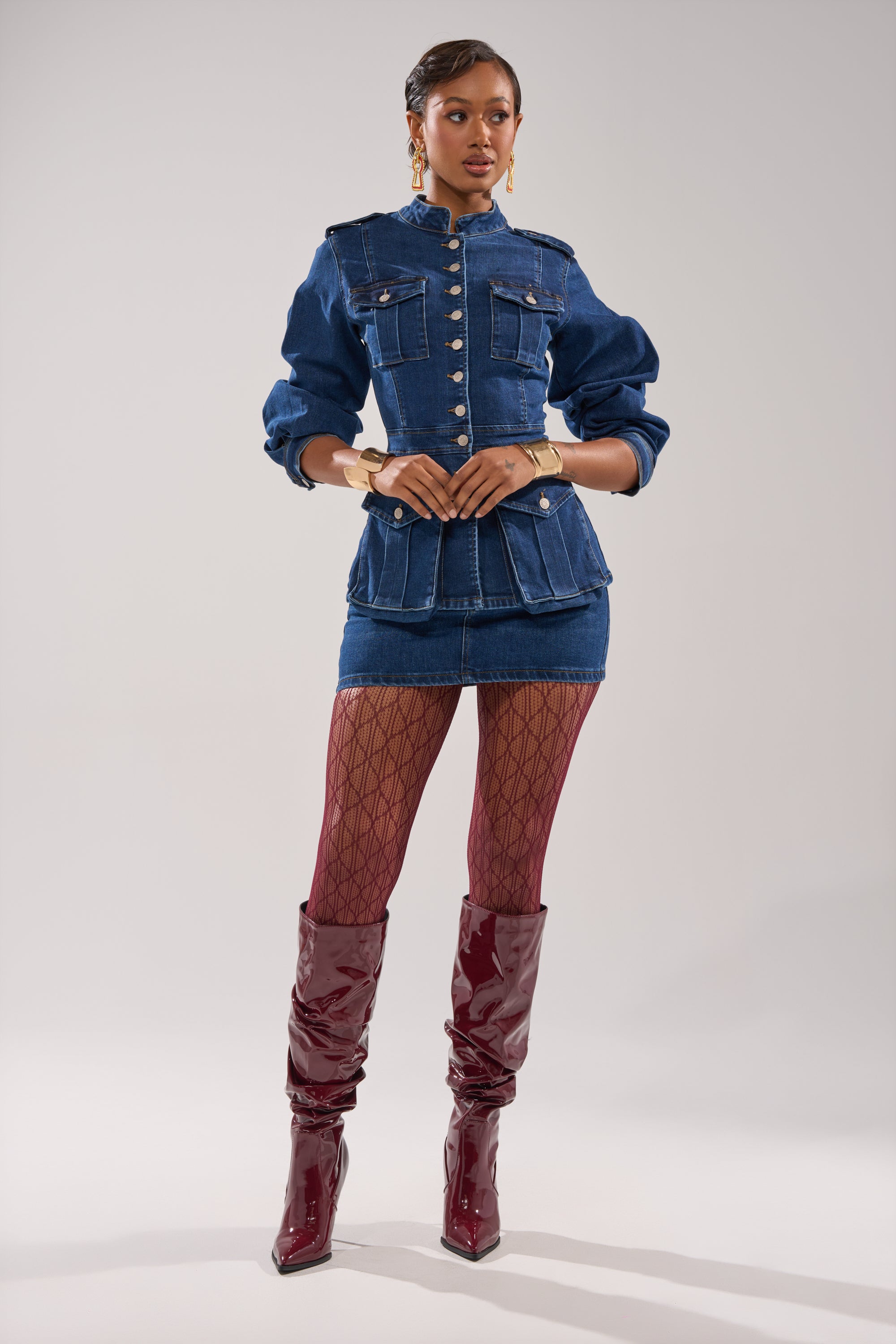 A woman stands confidently in the CRAZY FOR YOU LONG SLEEVE DENIM BUTTON DOWN and a matching mini skirt, styled with patterned fishnet tights, burgundy knee-high patent boots, gold hoop earrings, and a gold bracelet against a light background.