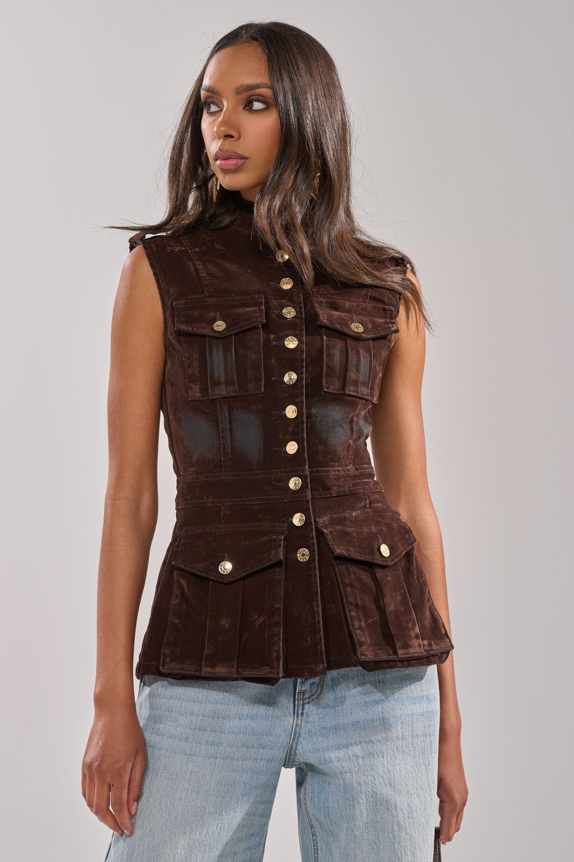 A woman wears the BACK WITH IT VELVET BUTTON DOWN, a fitted brown military-style vest with gold buttons and flap pockets, paired with light blue jeans against a plain light gray background.