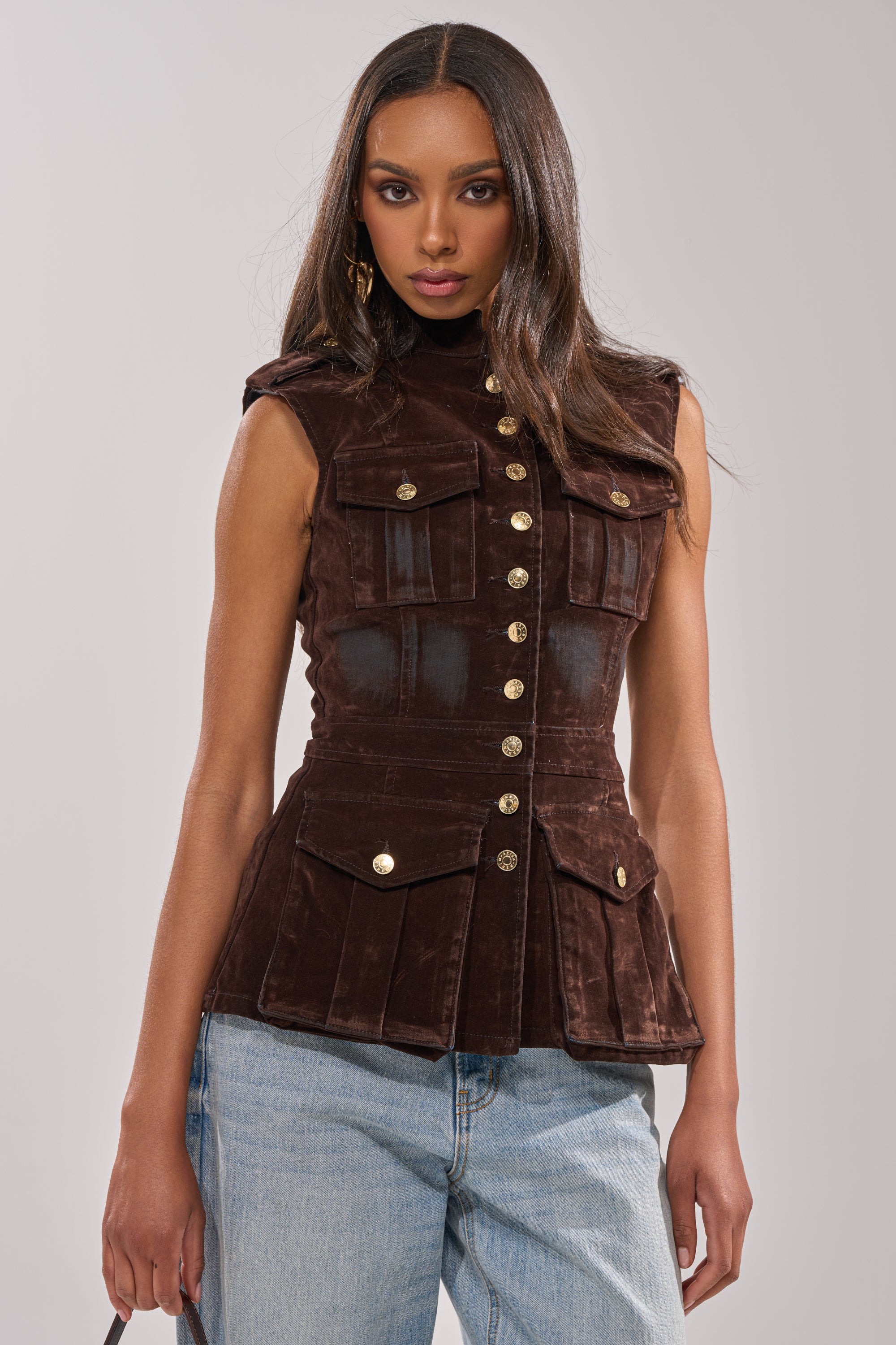 A woman with long brown hair wears the BACK WITH IT VELVET BUTTON DOWN, a fitted brown velvet vest with gold buttons and flap pockets, styled with light blue jeans against a plain light background.