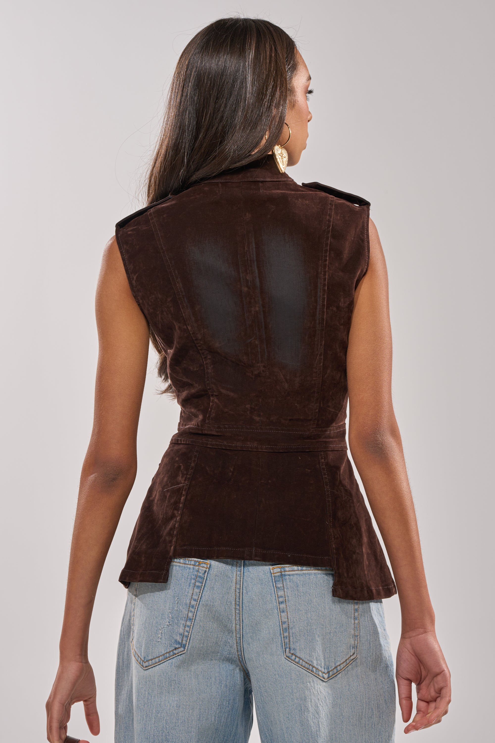 A person with long hair is seen from the back wearing the BACK WITH IT VELVET BUTTON DOWN, a fitted brown vest with a sheer panel, paired with light blue high-waisted jeans against a plain, light background.