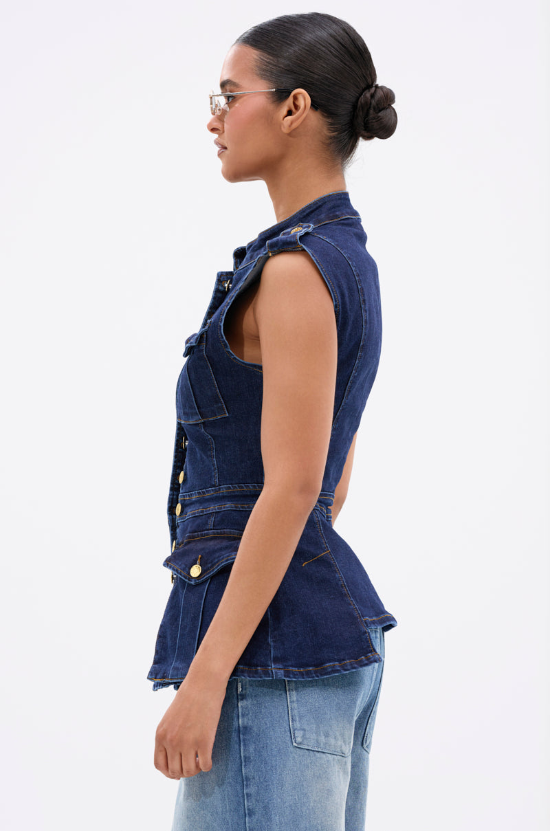 A woman with her hair in a bun and glasses wears the UMI STRETCH DENIM TOP in dark blue with gold hardware and light blue jeans, standing in profile against a plain white background.
