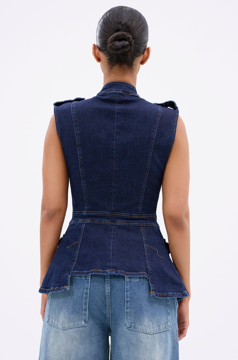 A person faces away, wearing the UMI STRETCH DENIM TOP—a dark blue, sleeveless vest with shiny gold hardware—paired with matching loose denim jeans. Their hair is in a neat bun against a plain, light background.