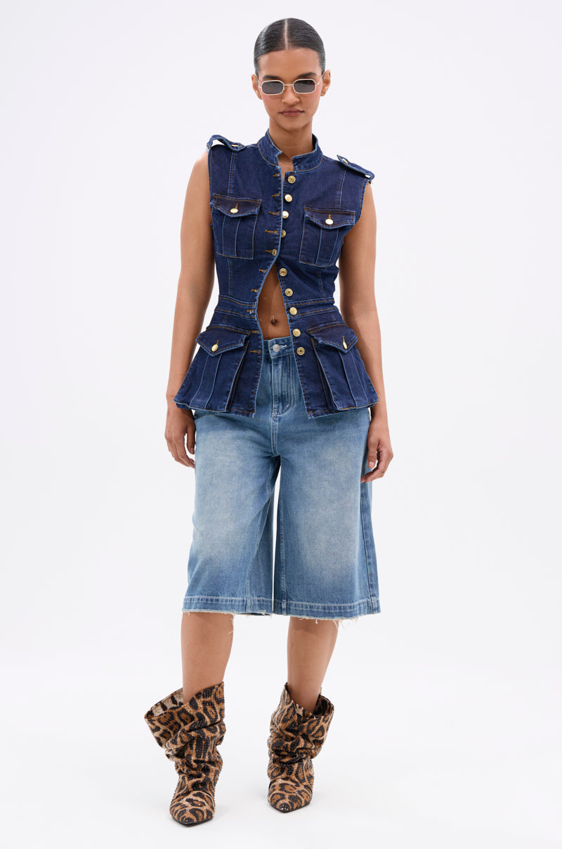 A woman faces forward in the UMI STRETCH DENIM TOP with high-shine gold hardware, paired with knee-length wide denim shorts, leopard print slouch boots, and sunglasses. Hair pulled back and a neutral expression complete her utility-chic look.
