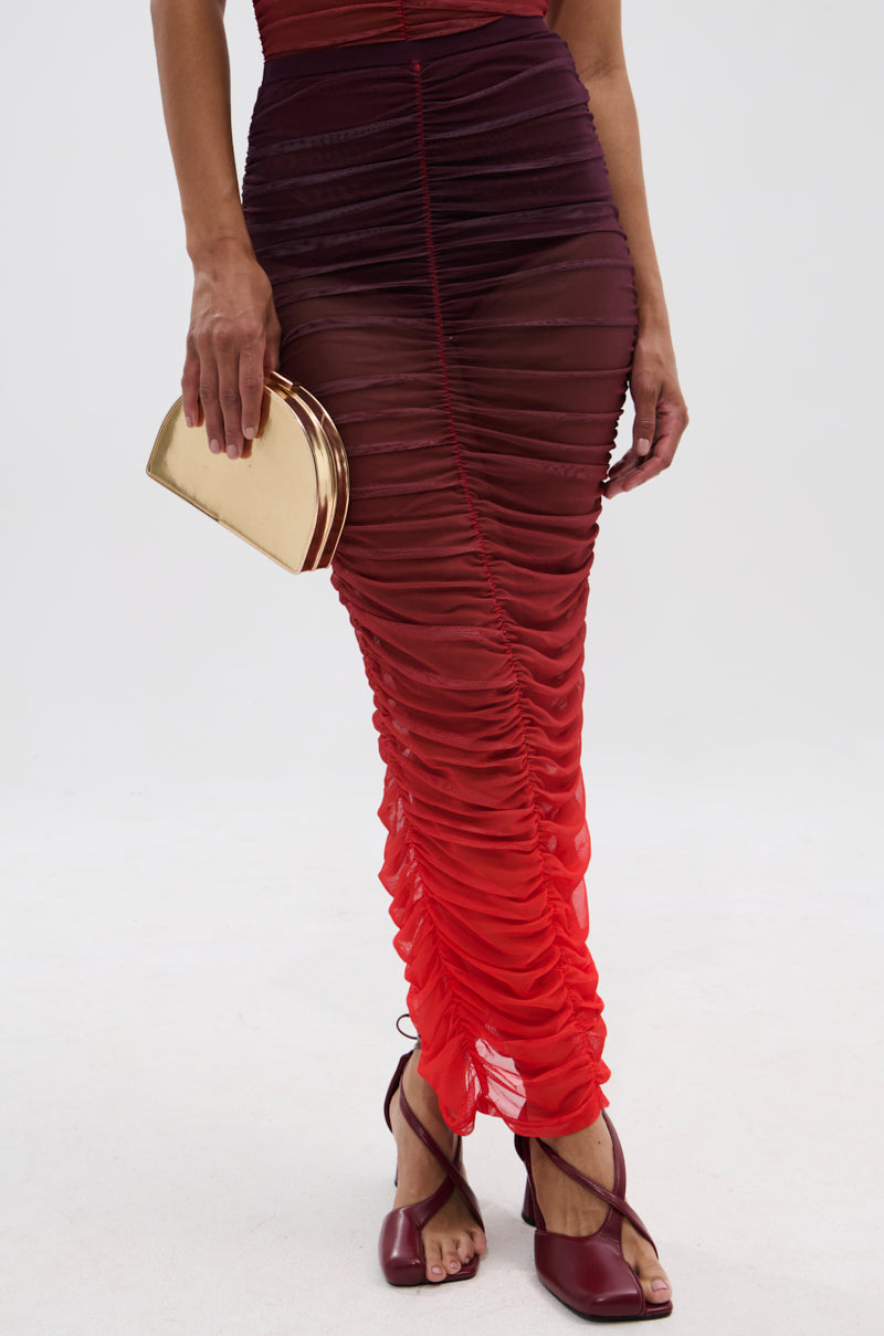 A woman wears the OH FIRE OMBRE MAXI SKIRT with ruched detailing, fading from dark burgundy to bright red, styled with a small gold clutch and burgundy heeled sandals against a plain white background.
