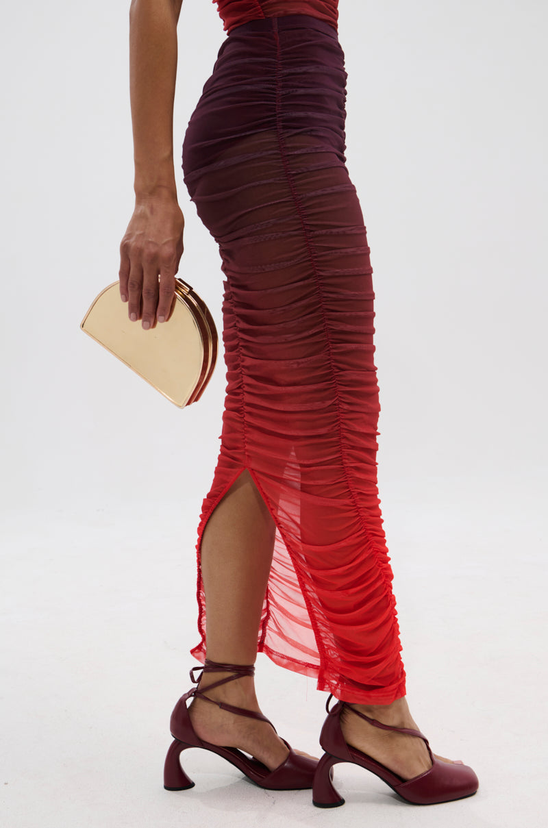 Wearing the OH FIRE OMBRE MAXI SKIRT with a fitted, ruched, sheer red and burgundy ombre design, paired with a small gold clutch and burgundy ankle-strap heels; set against a plain white background.