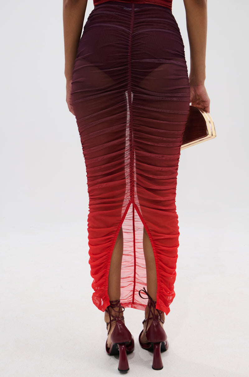 A person wears the OH FIRE OMBRE MAXI SKIRT—sheer, ruched red mesh with a back slit—paired with a small purse and lace-up high heels, photographed from behind against a plain background.