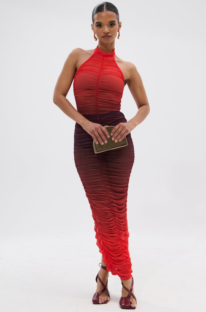 A woman poses against a plain white background, wearing the OH FIRE OMBRE MAXI SKIRT in a sleeveless style with red and burgundy gradient hues. She pairs it with burgundy sandals and holds a small brown clutch, one hand on her hip.