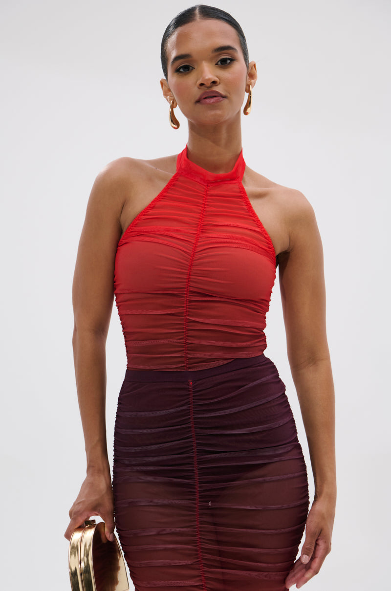 A woman wears the OH FIRE OMBRE MESH BODYSUIT, styled as a sleeveless ruched dress with a red halter top and sheer maroon skirt, holding a small gold clutch against a plain light background.