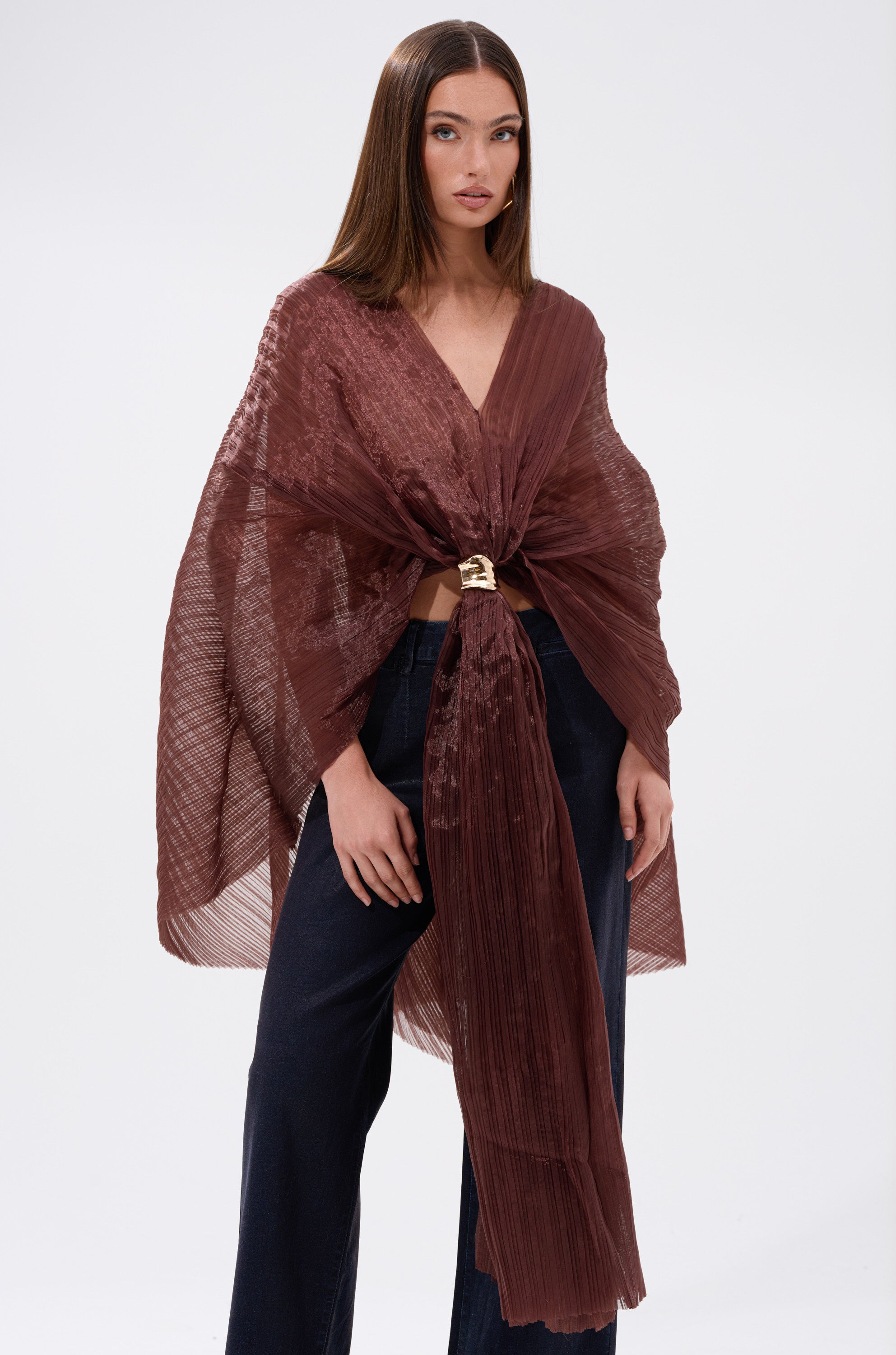 A woman with straight brown hair models the FLY AWAY WITH ME BLOUSE IN BRONZE— a sheer, pleated wrap top with batwing sleeves and a bow front tie — paired with dark wide-leg pants against a light background.