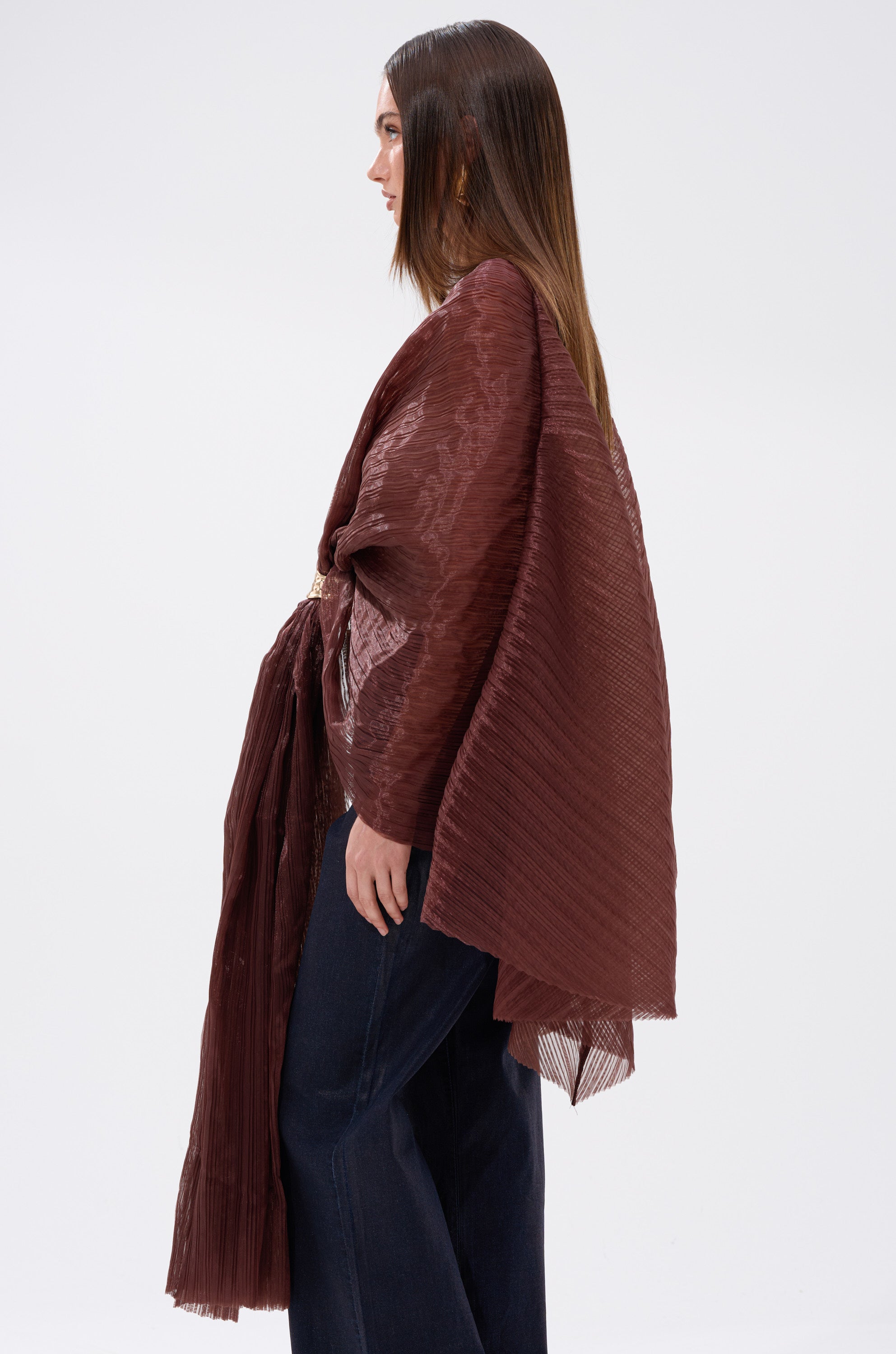 A woman stands in profile wearing the FLY AWAY WITH ME BLOUSE IN BRONZE, a textured, loose-fitting wrap top with batwing sleeves, paired with dark wide-leg pants. Her straight hair is center-parted against a plain white background.