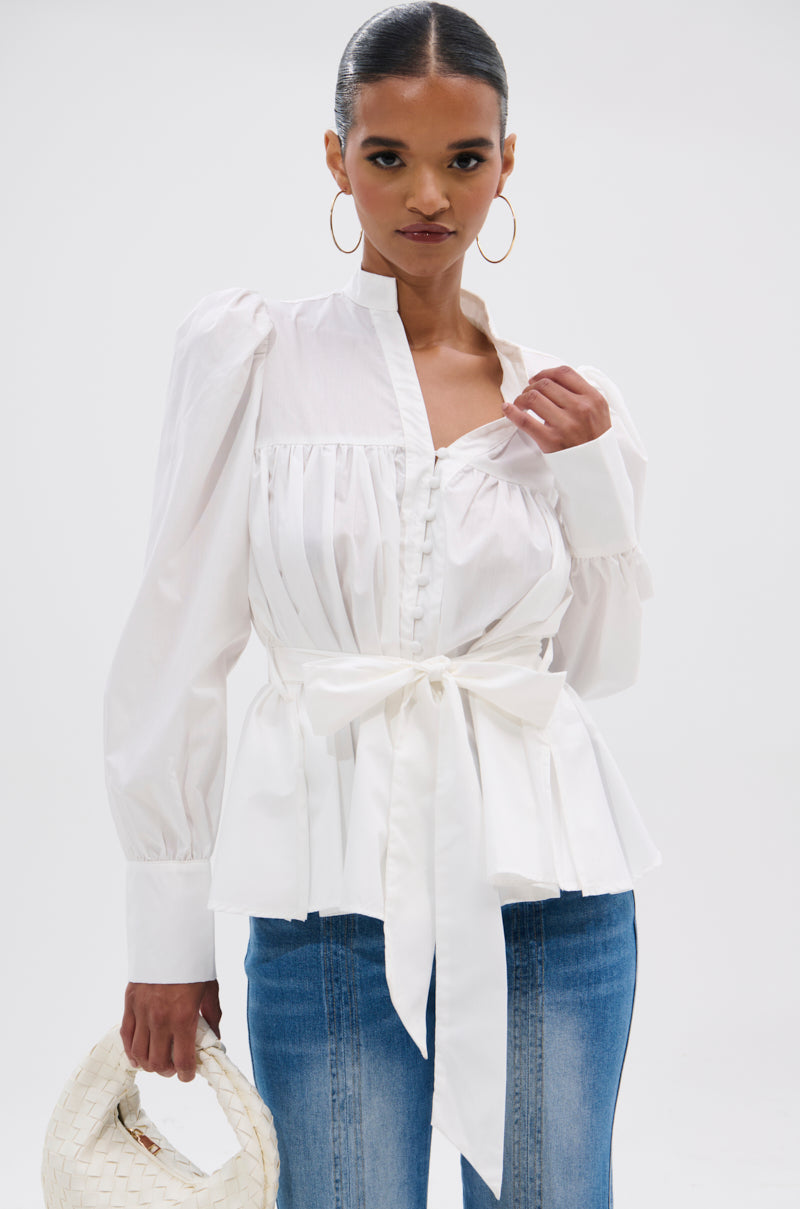 A woman in the LADY VIBES BUTTON DOWN BLOUSE WITH BELT, featuring puffed sleeves and a waist belt, paired with blue jeans and holding a white woven handbag, stands against a plain white background.