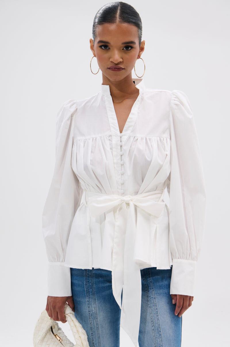 A woman styles the LADY VIBES BUTTON DOWN BLOUSE WITH BELT—a white, long sleeve, v-neck top with a waist tie—with blue jeans and a white woven clutch. Large hoop earrings and pulled-back hair complete her chic outfit.
