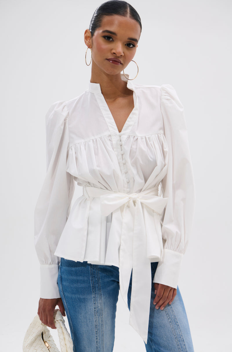 A woman wears the LADY VIBES BUTTON DOWN BLOUSE WITH BELT, styled with blue jeans and a white braided handbag. Her hair is pulled back, and she accessorizes with large hoop earrings.