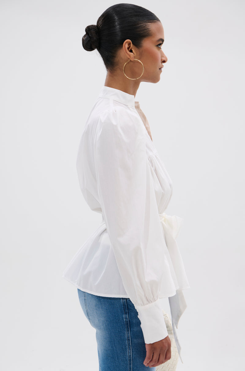 A woman with her hair in a neat bun, wearing the LADY VIBES BUTTON DOWN BLOUSE WITH BELT and blue jeans, stands in profile against a white background, accessorized with large hoop earrings.