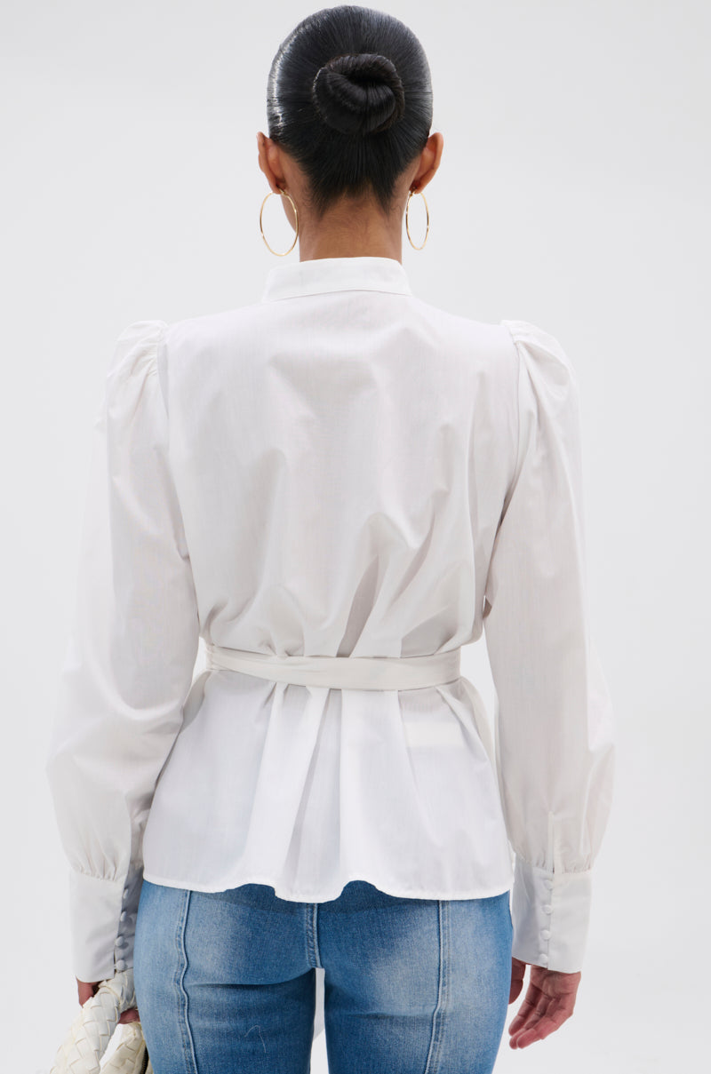 A woman, hair in a neat bun and wearing large hoop earrings, models the LADY VIBES BUTTON DOWN BLOUSE WITH BELT—featuring white cotton poplin fabric, puffed shoulders, and a belted waist—paired with blue jeans.