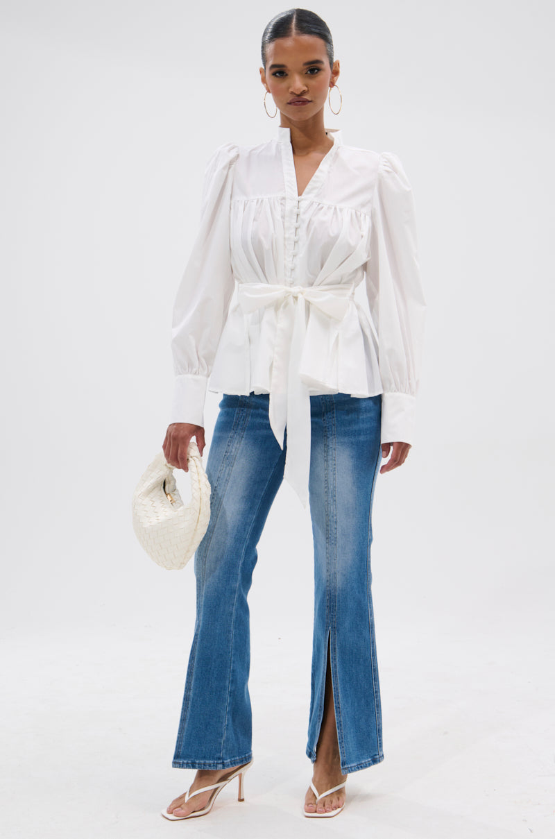 A woman stands against a white background wearing the LADY VIBES BUTTON DOWN BLOUSE WITH BELT, blue flared jeans with front slits, white heeled sandals, and holding a white woven handbag.