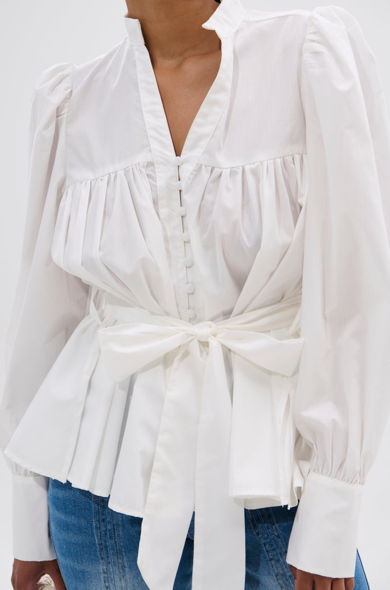A person wears the LADY VIBES BUTTON DOWN BLOUSE WITH BELT, featuring puffed shoulders, a V-neckline with fabric-covered buttons, gathered details, and a wide sash at the waist, paired with blue jeans.