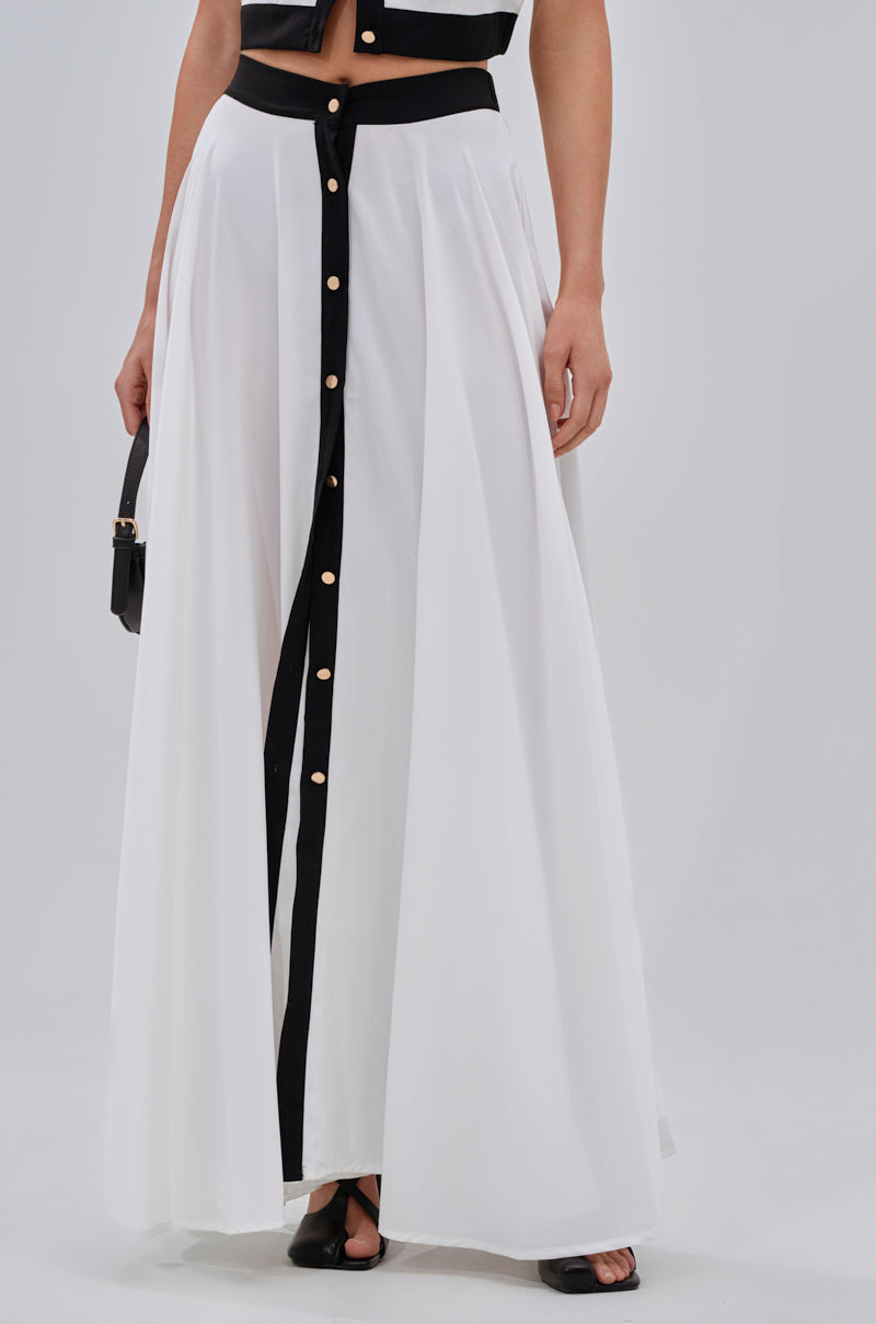 A person wearing the VIVI MAXI SKIRT, a silky chiffon maxi with black vertical trim and gold front buttons, styled with black open-toe shoes and a small black handbag. 1