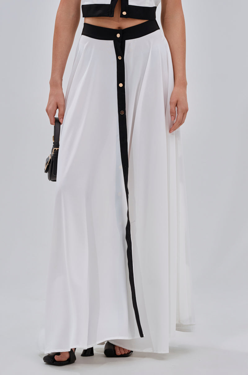 Wearing the VIVI MAXI SKIRT, a flowing white A-line maxi with black trim and buttons, paired with a cropped black top, black handbag, and black sandals for a complete look. 2