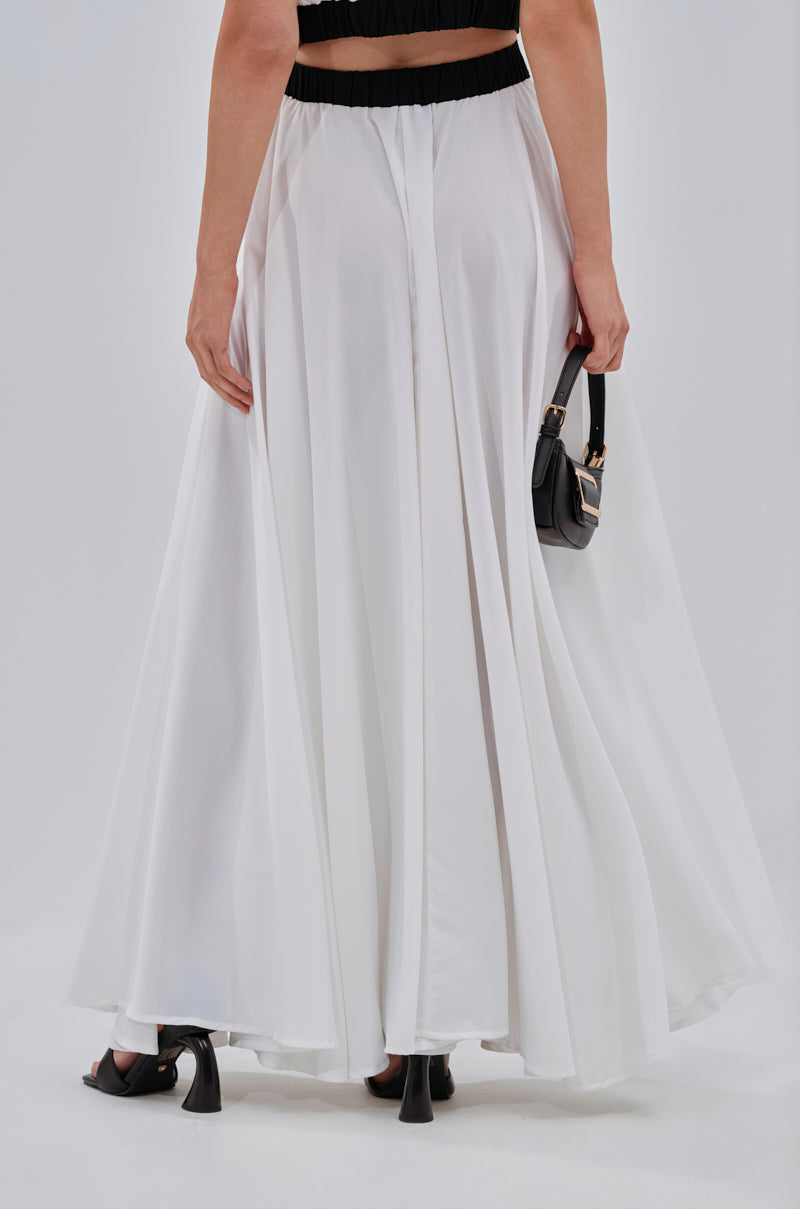 A woman is photographed from behind wearing the VIVI MAXI SKIRT—a flowing white chiffon A-line maxi—paired with black heels and a small black handbag, set against a plain light background. 4