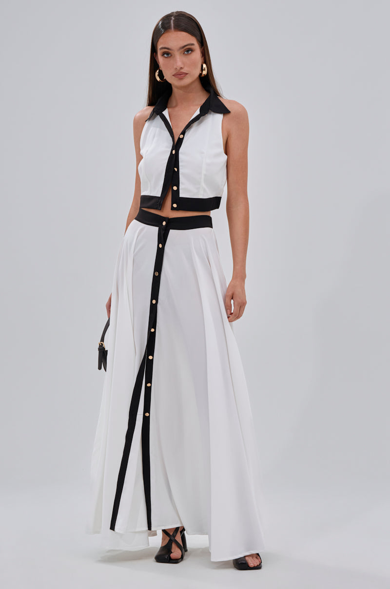 A woman wears a sleeveless white two-piece with black collar, button accents, and black-trimmed panels. The silky chiffon set features a cropped top and the VIVI MAXI SKIRT in an A-line silhouette. She carries a small purse and wears black heels. 5