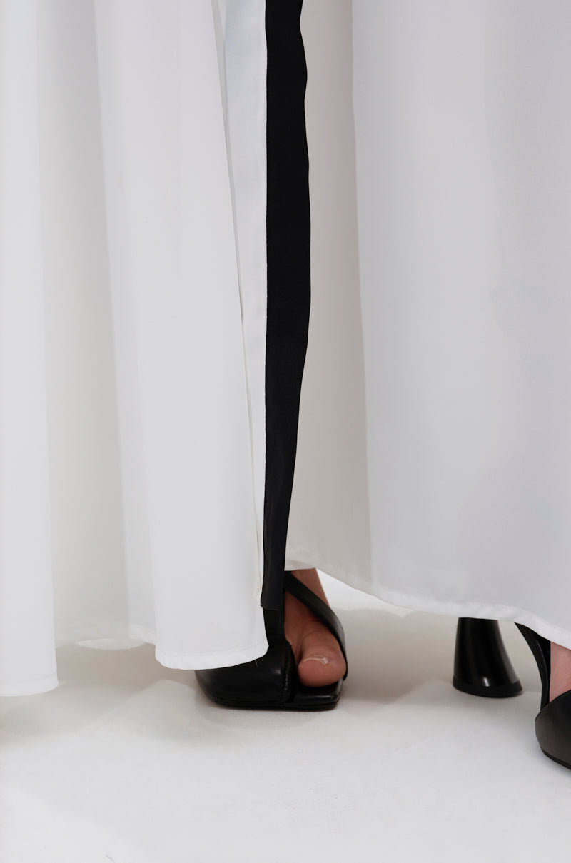 Close-up of the lower part of the VIVI MAXI SKIRT, a white flowy maxi skirt with a black vertical stripe, shown with black open-toed heeled shoes. The background is white. 7