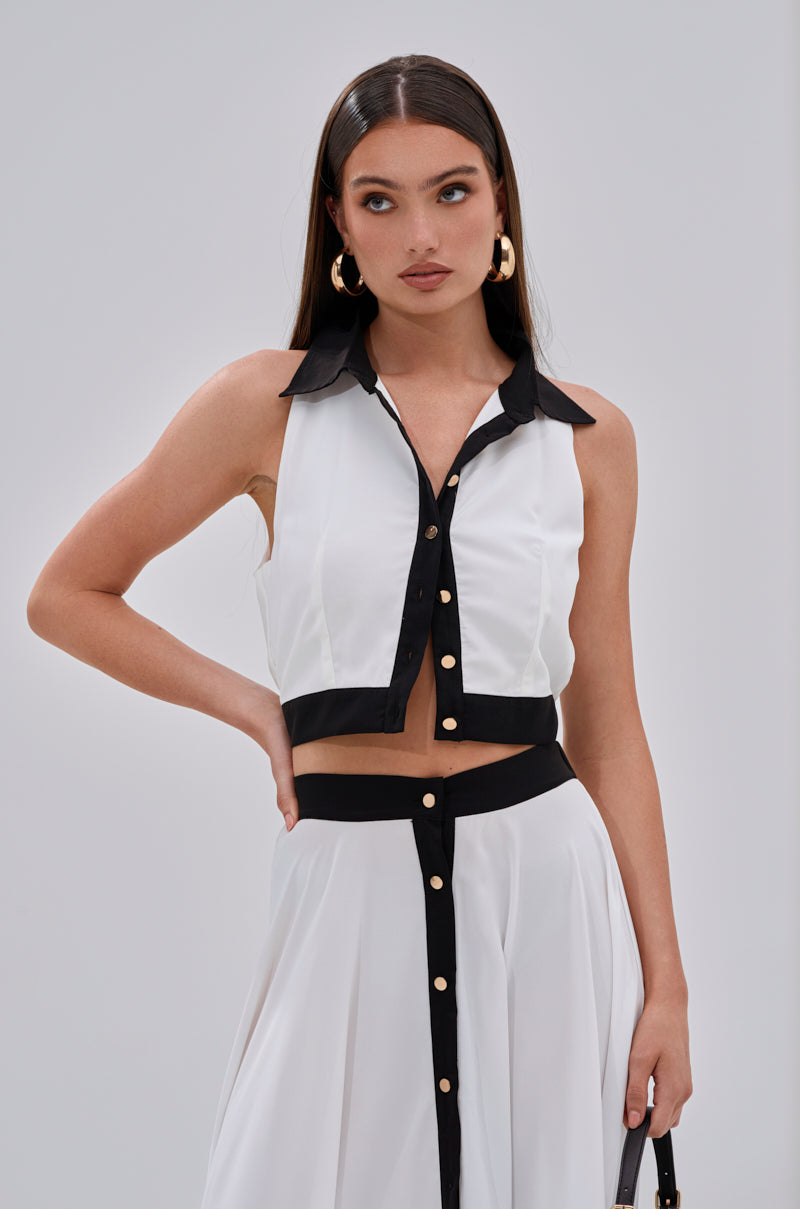 A woman models the VIVI BUTTON DOWN BLOUSE, a sleeveless white cropped top with a black collar and trim, paired with a high-waisted skirt with black detailing and gold buttons. She has long straight hair and wears large hoop earrings. 0