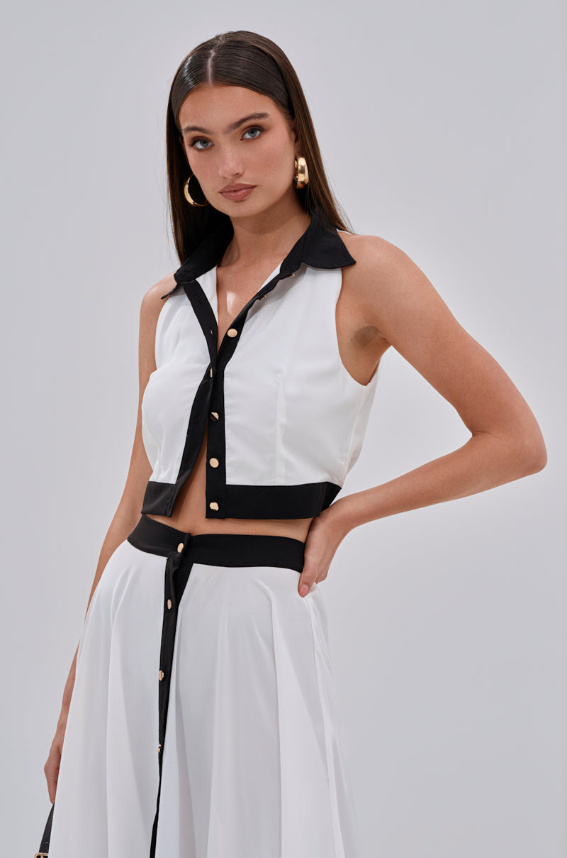 A woman with long brown hair models the VIVI BUTTON DOWN BLOUSE, a cropped white button-down top with black collar, trim, and gold buttons, paired with a matching skirt featuring a black waistband against a light gray background. 1