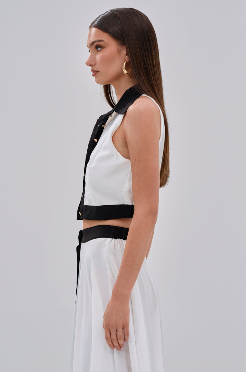 A woman with straight brown hair wears the VIVI BUTTON DOWN BLOUSE, a sleeveless chiffon crop top with black accents, and a matching white skirt, standing in profile against a plain light grey background. 2