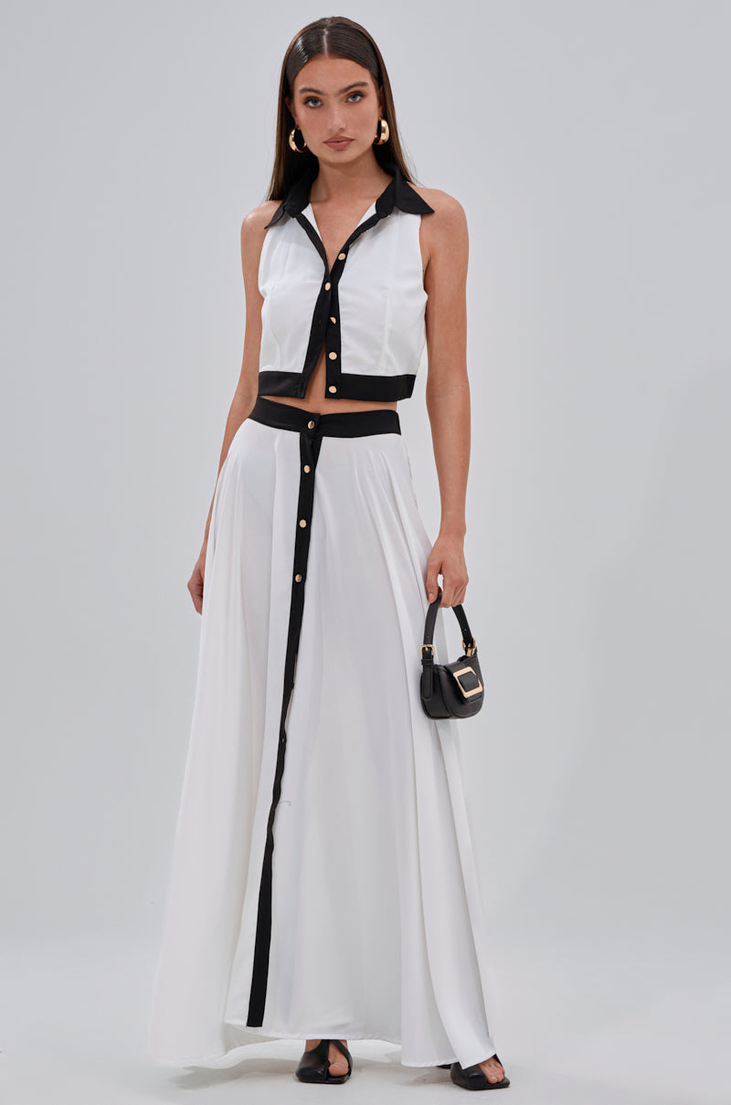 A woman wears the VIVI BUTTON DOWN BLOUSE, a sleeveless black and white cropped top, with a matching long skirt, black sandals, gold earrings, and a small black handbag, standing against a plain light gray background. 5