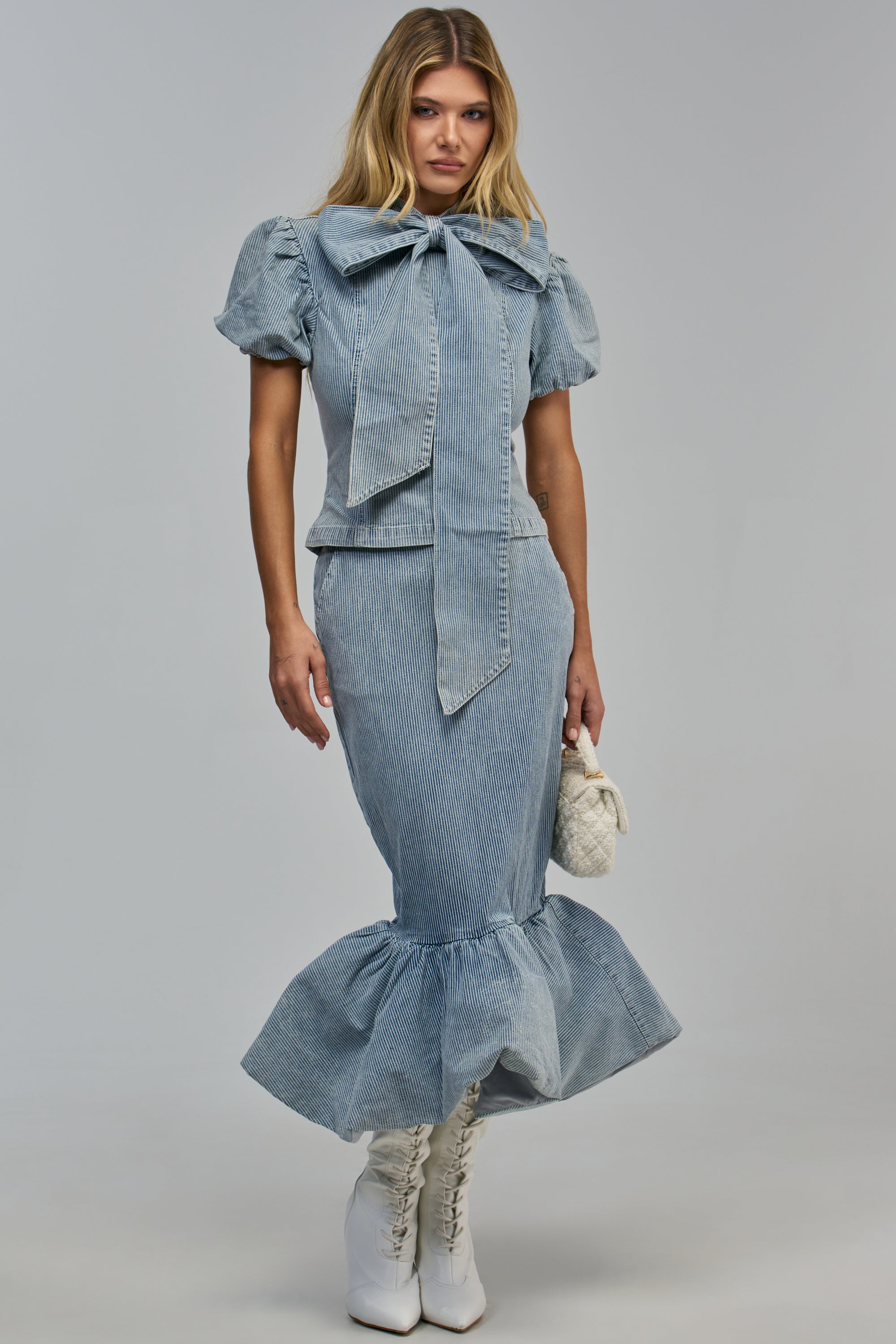 A person wears a light blue pinstriped denim puff-sleeve top with a pussycat bow and the DENIM DEBUT MIDI SKIRT with a ruffled hem, paired with white boots and a small white handbag.