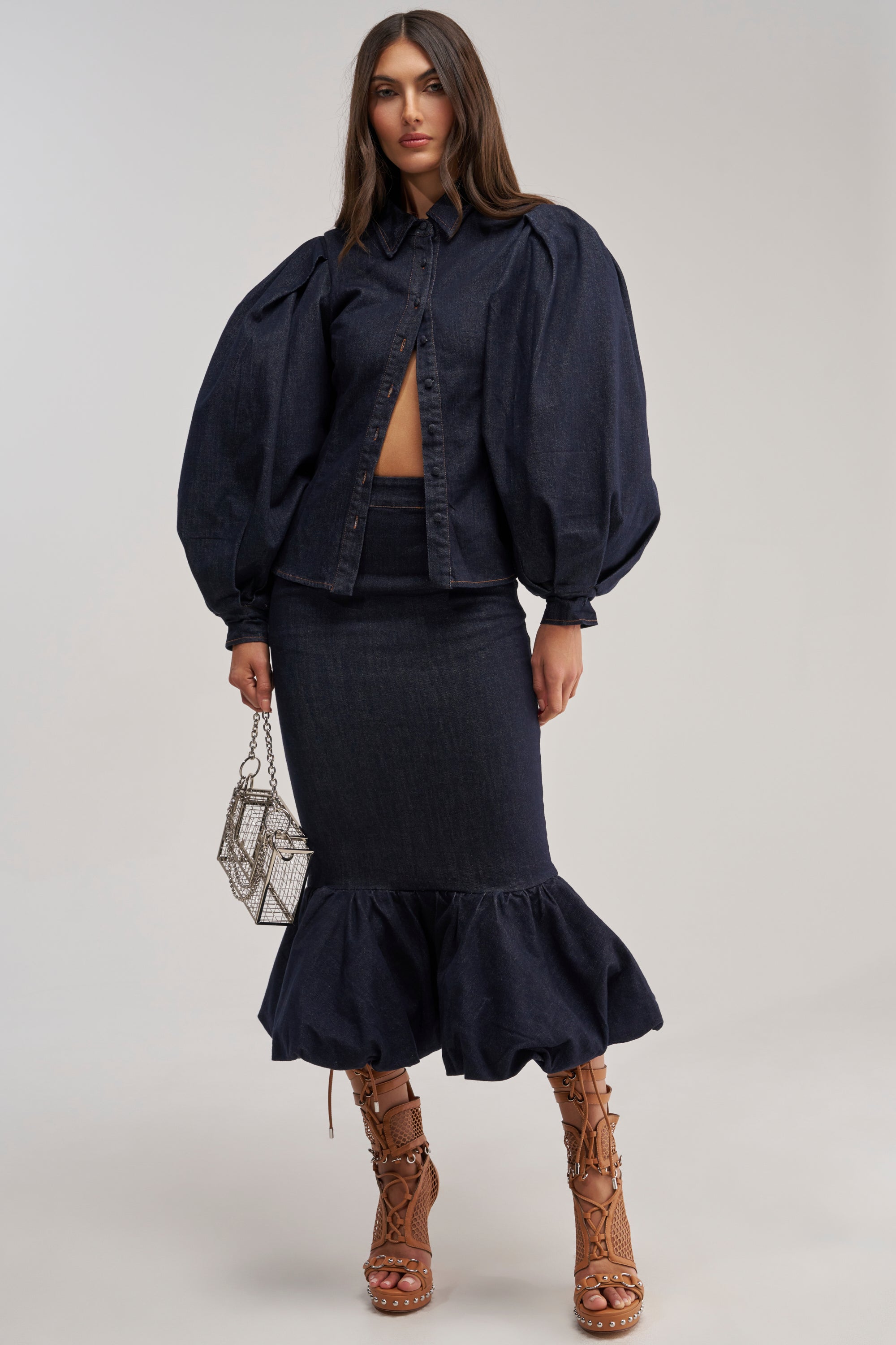 A woman wears the LET'S CHANGE IT UP DENIM MIDI SKIRT, paired with a dark button-up top with puffed sleeves, a silver mesh bag, and tan lace-up high-heeled sandals.