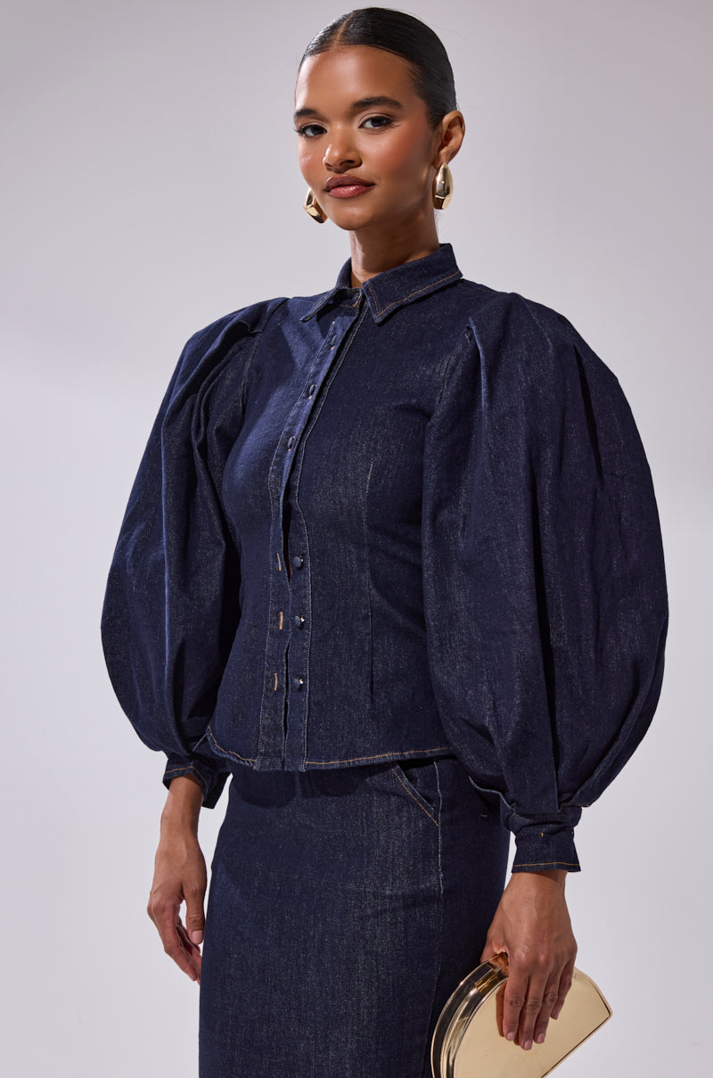 A woman wearing the LET'S CHANGE IT UP DENIM BLOUSE and a matching dark-wash skirt, holding a small beige clutch, stands against a plain light background.