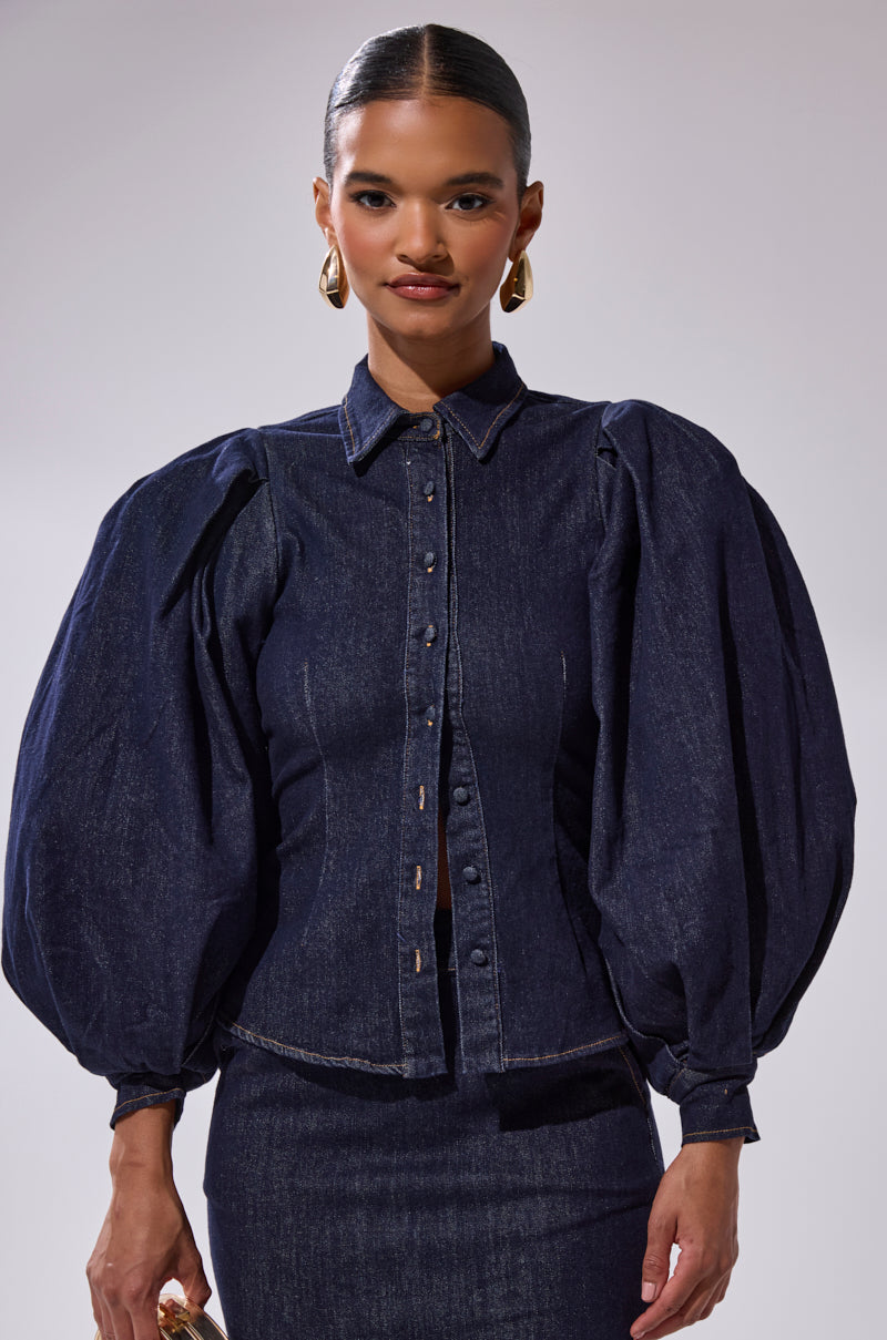 A woman wears the LET'S CHANGE IT UP DENIM BLOUSE, featuring dark-wash denim and dramatic balloon sleeves, paired with a matching skirt and large gold hoop earrings against a plain background.