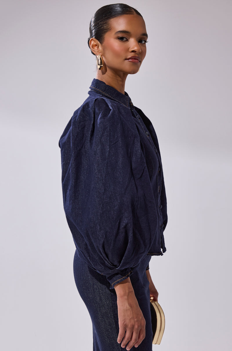 A woman models the LET'S CHANGE IT UP DENIM BLOUSE and matching skirt with balloon sleeves, accessorized with gold hoop earrings and a light clutch. Her hair is slicked back, and she poses sideways against a plain light background.