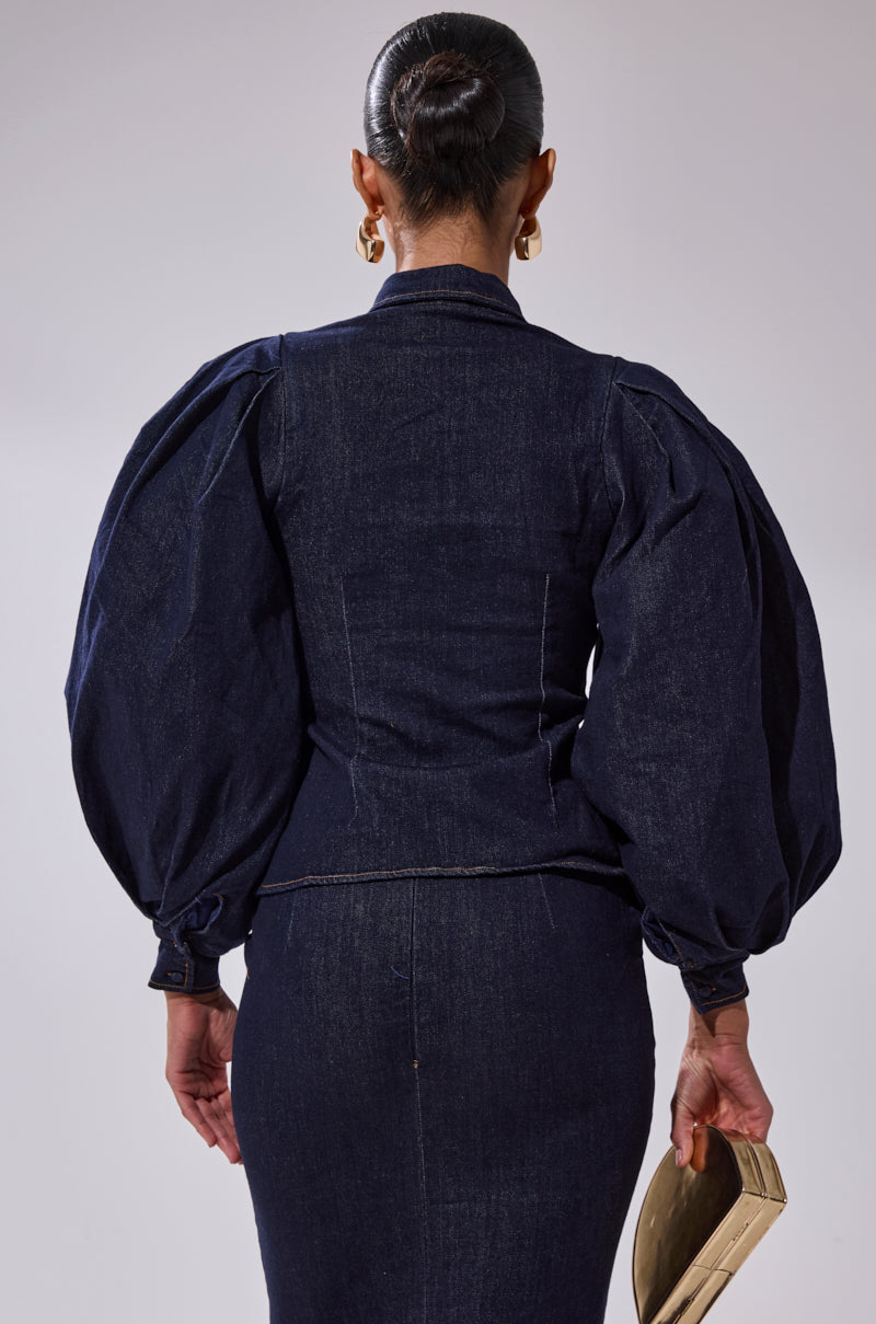 A person with hair in a neat bun, seen from the back against a plain light background, wears the LET'S CHANGE IT UP DENIM BLOUSE with dramatic balloon sleeves and holds a beige clutch.