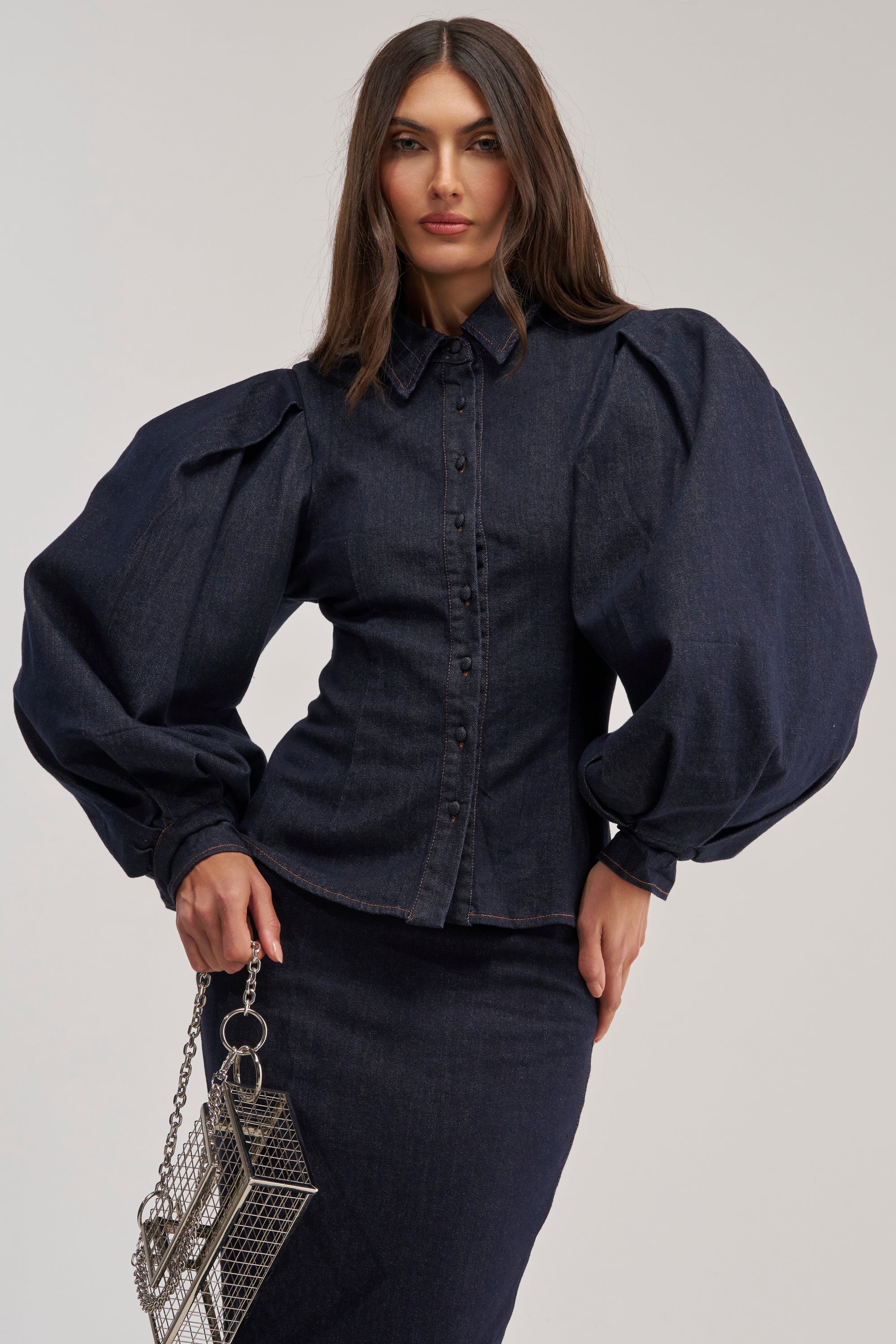 Wearing the LET'S CHANGE IT UP DENIM BLOUSE with bold balloon sleeves and a matching skirt, a woman holds a metallic geometric purse against a plain light background.