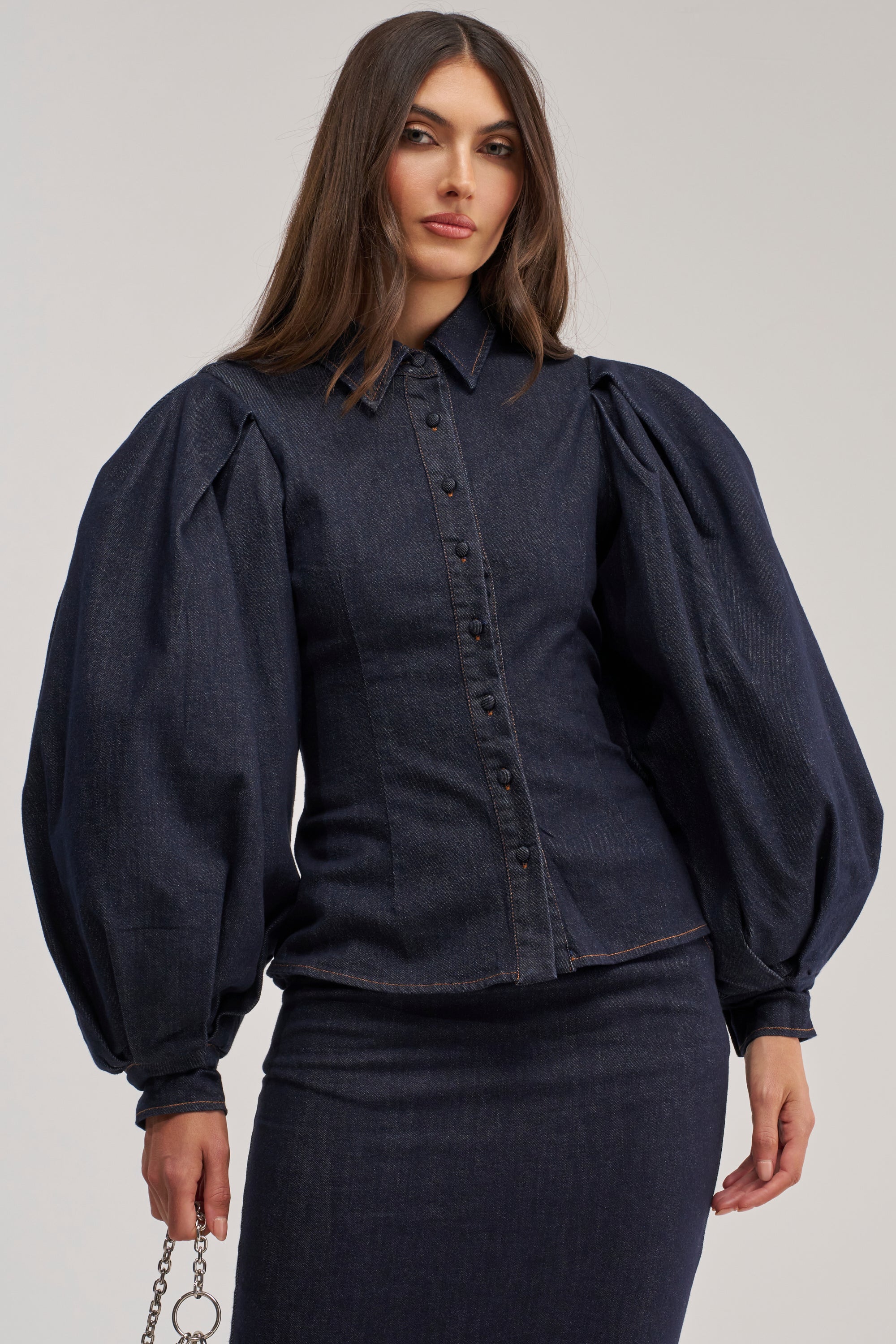 A woman models the LET'S CHANGE IT UP DENIM BLOUSE with dramatic balloon sleeves and a matching fitted skirt, standing against a plain light background, holding a chain accessory.