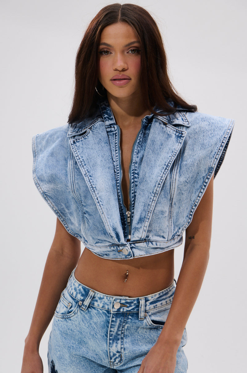 A woman with long brown hair wears the TOO COOL FOR YOU DENIM ZIP-UP BLOUSE, a sleeveless cropped top with wide shoulders, styled with high-waisted jeans for an edgy streetwear look against a plain light background. 2